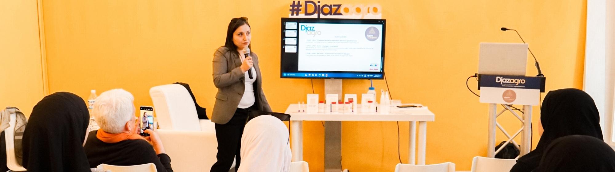 A speaker presents during a conference at the Djazagro trade show, addressing an attentive audience. A screen behind her displays the event program.