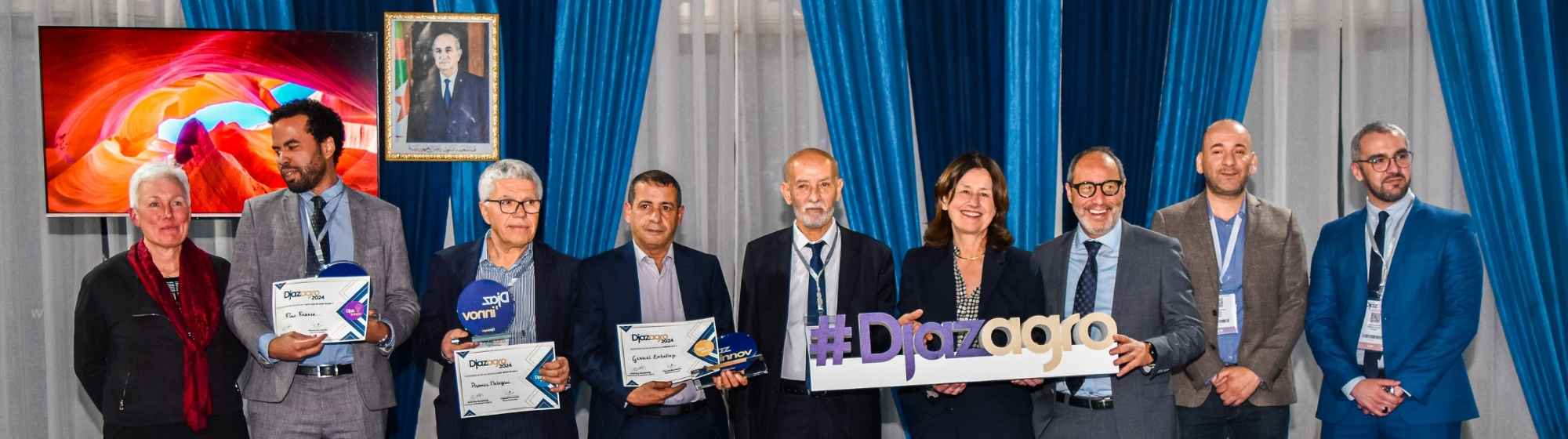 Groupe de lauréats et organisateurs à Djazagro 2024 posant avec des certificats et un panneau #Djazagro, dans un cadre formel avec des rideaux bleus et un portrait encadré en arrière-plan