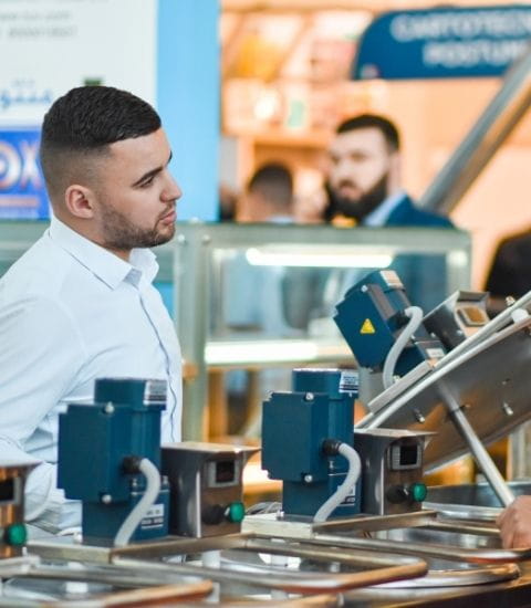 A man in a white shirt talks with an exhibitor in front of industrial equipment at the Djazagro trade show. In the background, other visitors and exhibition stands.