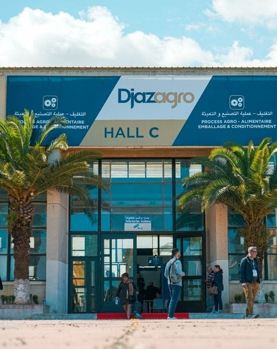 Entrée du salon Djazagro avec un panneau indiquant "Hall C", entourée de palmiers et de visiteurs entrant et sortant du bâtiment.