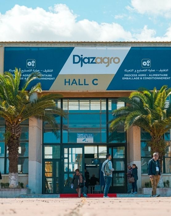 Entrance of the Djazagro trade show with a sign displaying "Hall C", surrounded by palm trees and visitors entering and exiting the building.