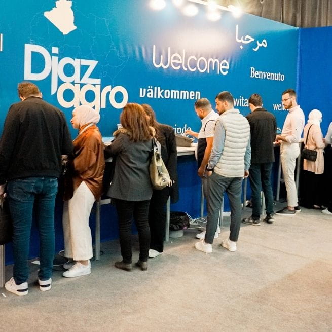 Visitors at a reception desk at the Djazagro exhibition, with a blue sign displaying welcome messages in multiple languages